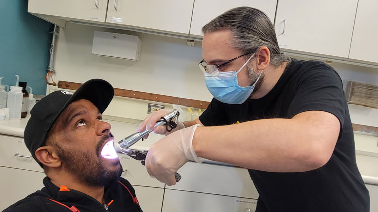 Close-up of two people posing as dentist and patient, one wearing surgical mask and holding flashlight and another tool while peering into the other’s open mouth.
