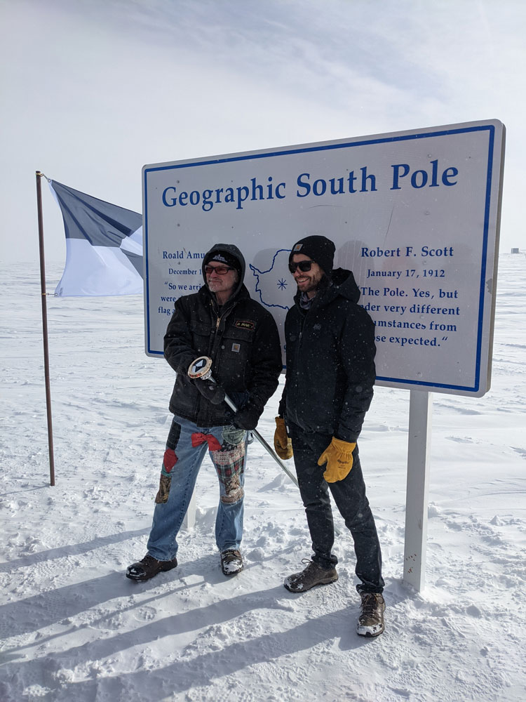 Two people standing in front of geographic South Pole sign and holding the pole marker.