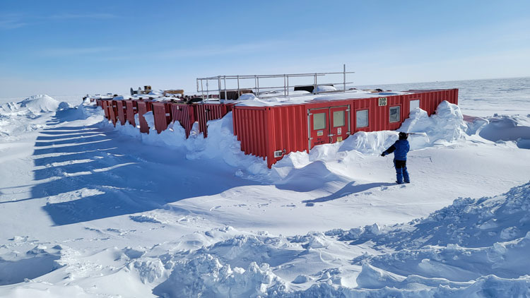 Person in parka standing near large shipping containers out on the ice at the South Pole.