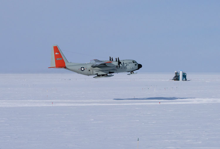 Side view of Hercules plane on lift off at the South Pole, with the IceCube Lab building small in the background.