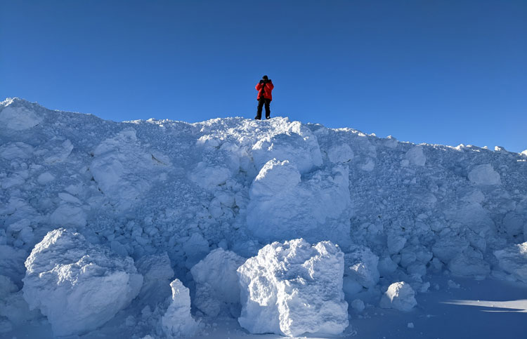 Person standing at edge of snow wall that is about four times their height.