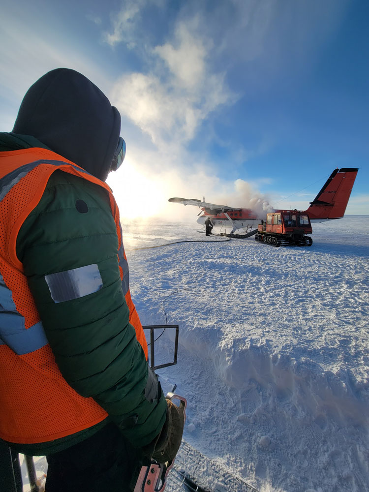 South Pole worker from behind, in foreground, watching plane refueling in distance.