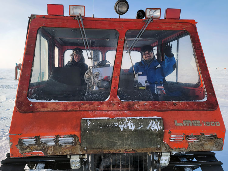 Front view of red snow vehicle, with two winterovers inside seen through windshield, smiling and waving to camera. 