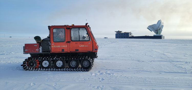 Side view of red snow vehicle, with South Pole telescope in distance.