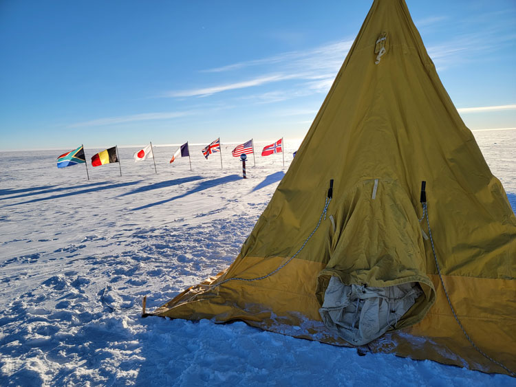A Scott tent pitched on the ice at the South Pole, with the ceremonial Pole marker and some flags seen in the background.