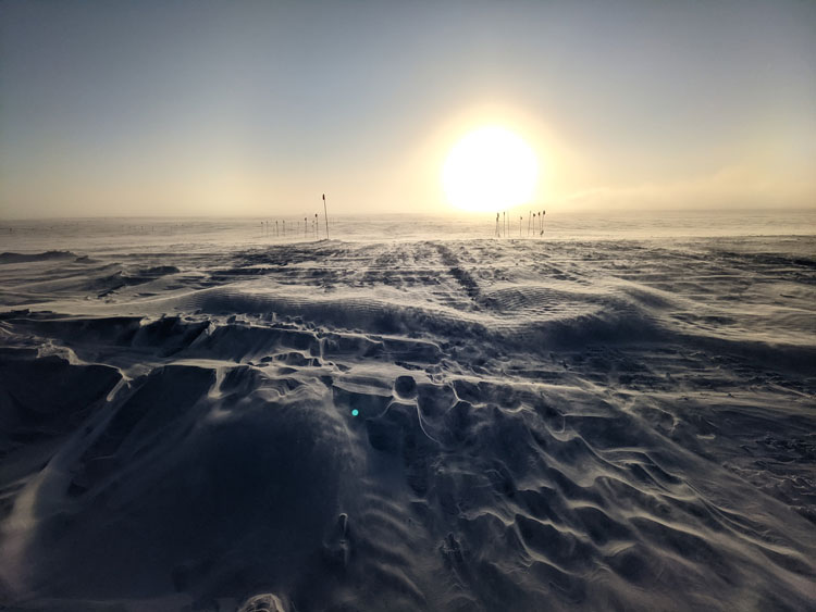 Bright sun low on horizon at the South Pole, with a few flags visible in the distance.