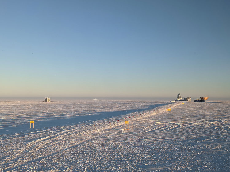 Long shadows cast on the icy surface at the South Pole from buildings in the Dark Sector, off in the distance.