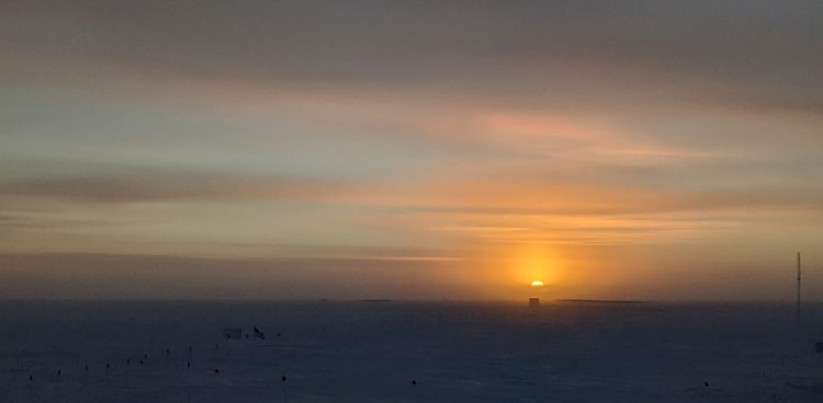 Orange sun low in sky at the South Pole, some thin horizontal clouds in the sky.