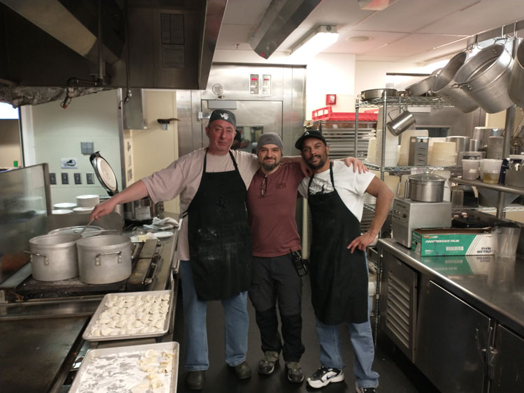 Three cooks in the South Pole kitchen, posing with arms around each other, with large pots and trays of dumplings next to the stove.