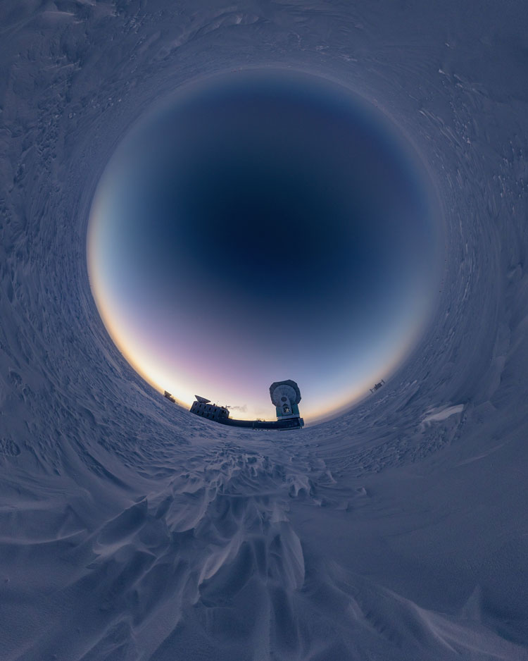 Circular panoramic view of snow and sunset at the South Pole, with South Pole Telescope in the foreground and the IceCube Lab in the distance.