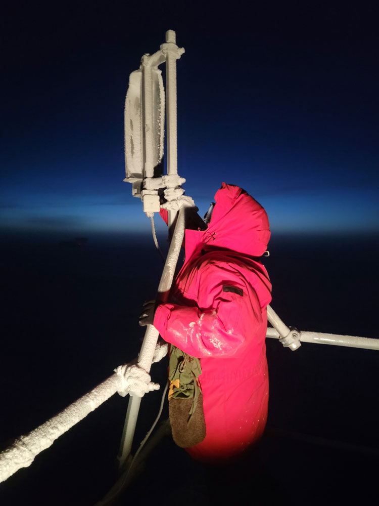Winterover in red parka checking equipment on roof of IceCube Lab at night.