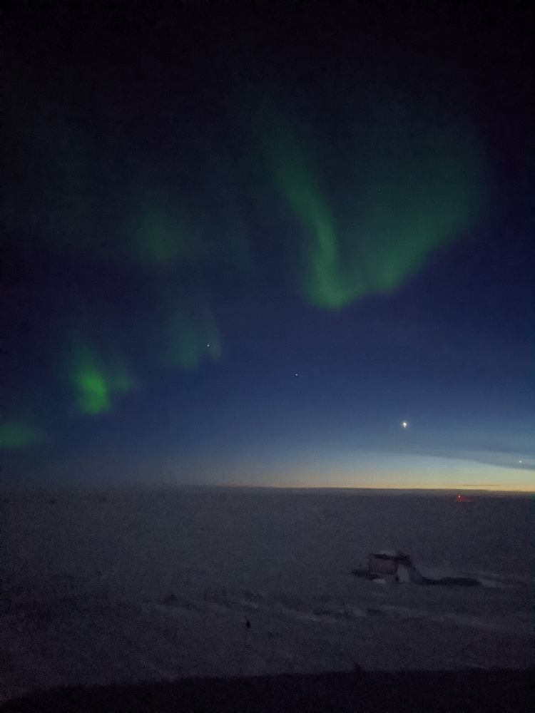 Night sky at the South Pole post sunset, with some light lingering along horizon, a bright planetary conjunction, and light wispy auroras overhead. 