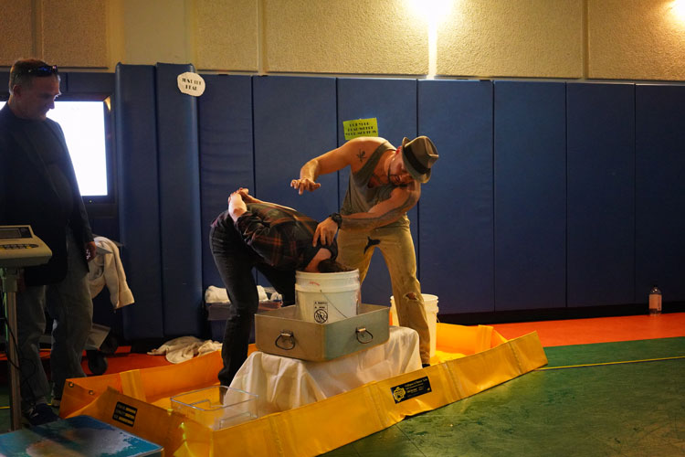 A person standing, dunking the head of a person kneeling into a large bucket of water
