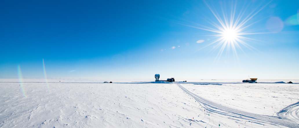 Bright sun in blue sky with several South Pole telescope buildings in distance along horizon