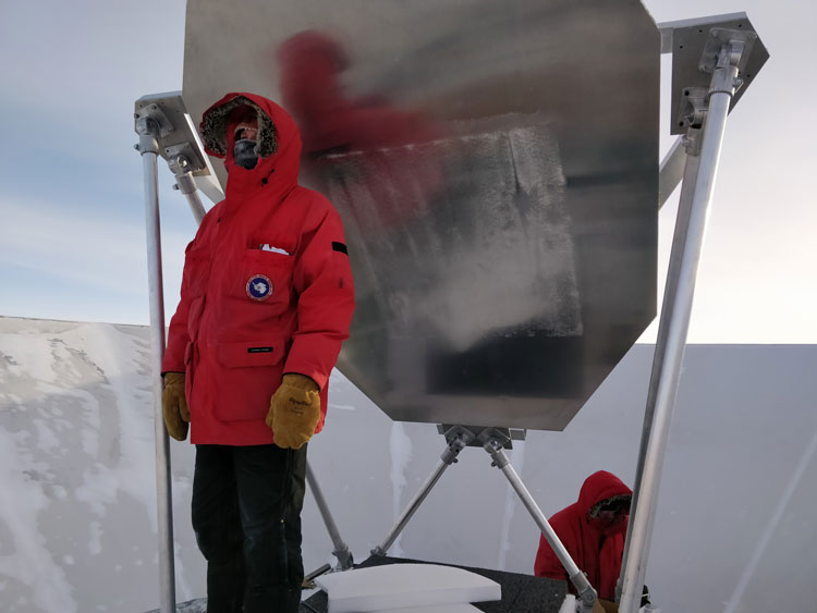 Person in red parka, standing near BICEP telescope, looking off into distance