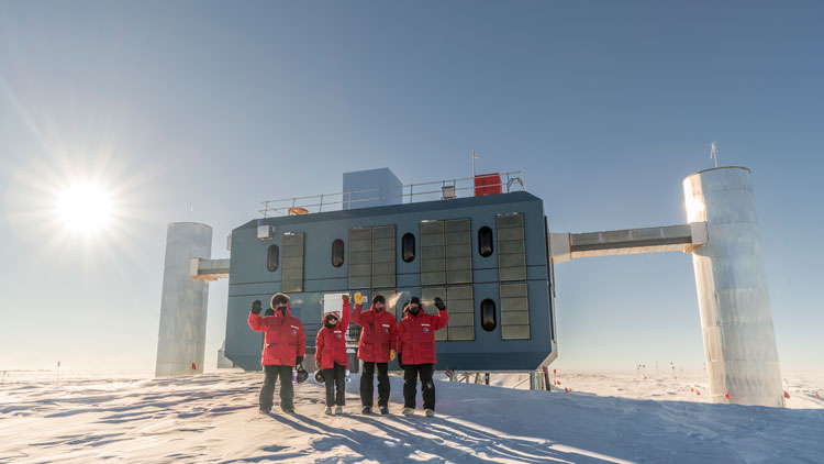 Four winterovers in red parkas waving outside the IceCube Lab