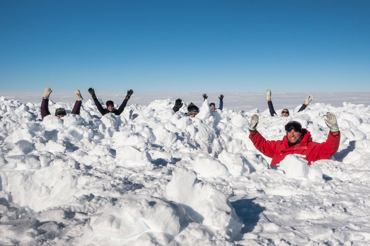 People with arms up in air, standing in chest-high holes dug into snow 