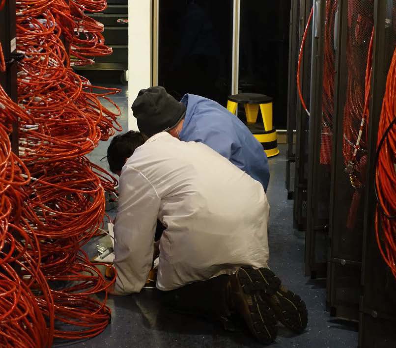 Two people from behind, hunched over in computer room with lots of visible cables