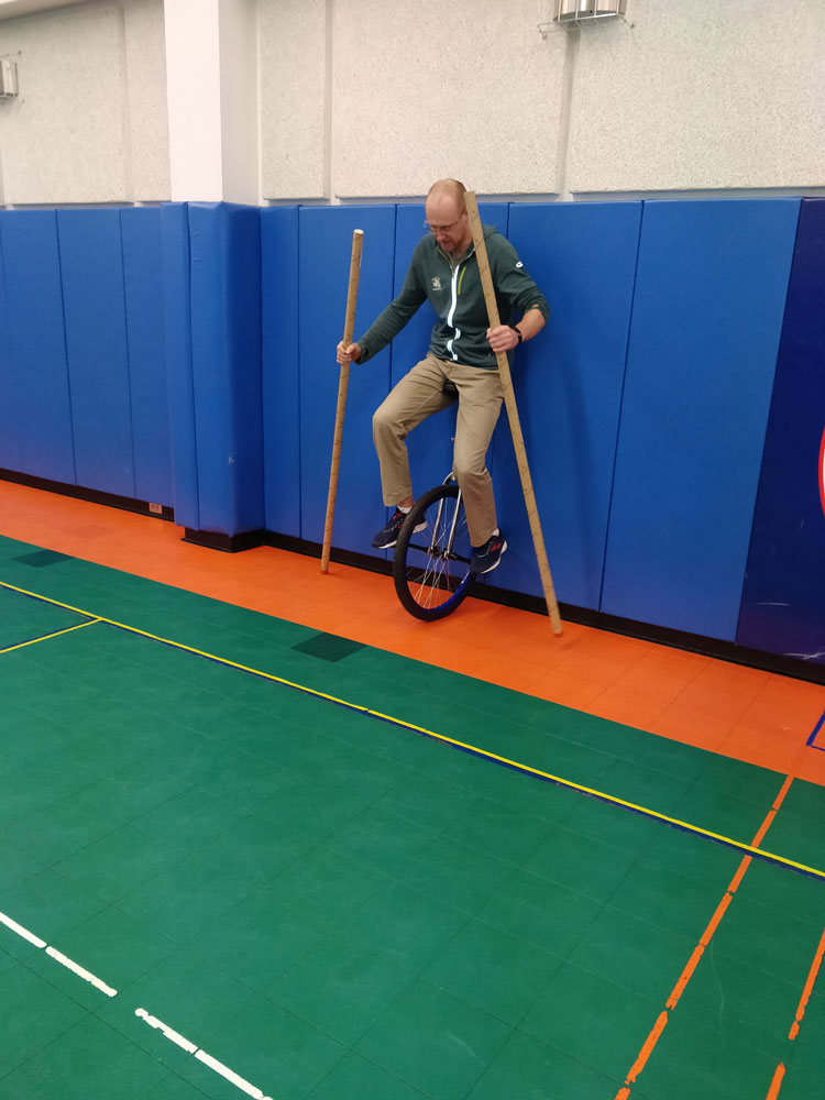 Person on unicycle, leaning back against padded wall and holding long balancing poles