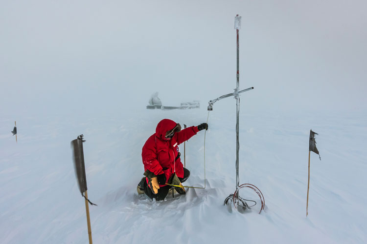 Person in red parka kneeling outside at south pole taking measurements