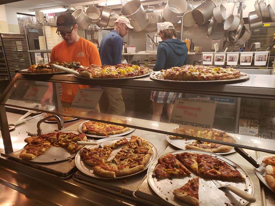 Trays of pizzas on kitchen warming shelves, with several people in kitchen in background