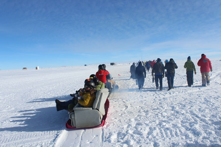 Sofa with people on it being pulled alongside runners in race at the south pole