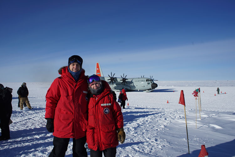 Two people in red parkas standing out on the ice with plane behind them