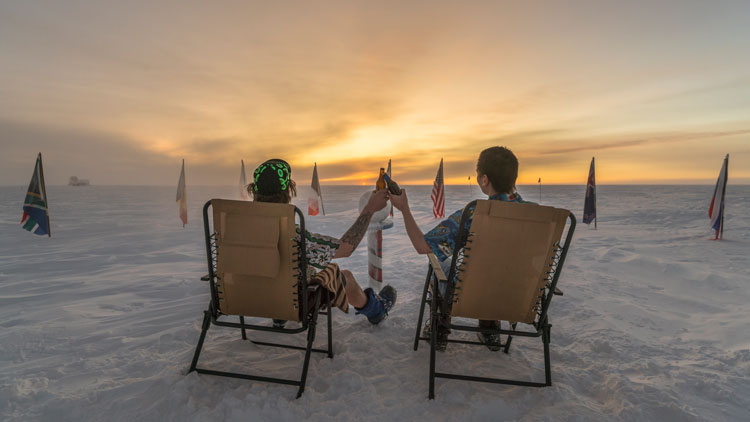 Two people seated, backs to camera, facing sunrise at South Pole