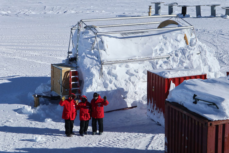Three people in red parkas, waving in distance near snow-covered large equipment