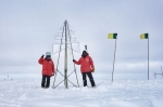 Two people in red parkas standing opposite sides of a large radio antenna